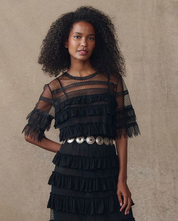 A woman with curly hair models THE GREAT. H25 COLLECTION's Winter Mesh Flounce Top in black, featuring sheer ruffles and layered details, styled with a metallic belt against a neutral background.