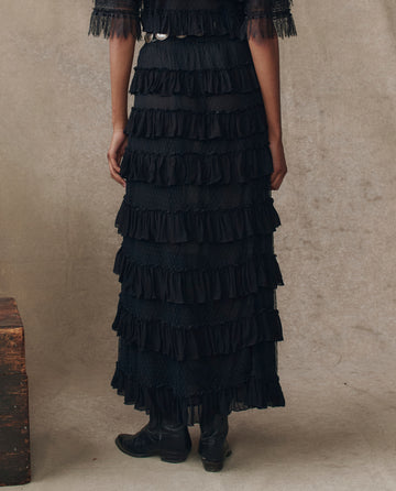 A person models THE GREAT. H25 COLLECTION’s Winter Mesh Flounce Skirt in black with black boots, standing against a neutral textured background.