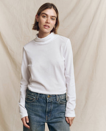 An elegant person in THE GREAT. HOL 24 TURTLENECKS' "The Turtleneck" in True White and timeless blue jeans poses confidently against a soothing beige backdrop.