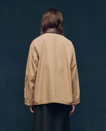 Rear view of a person with long hair wearing a light brown jacket and dark skirt against a dark background.