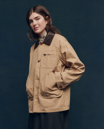 Against a dark, plain background, a person showcases The Barn Jacket by THE GREAT. SP25 COLLECTORS—a golden outerwear piece featuring large pockets and a dark collar.