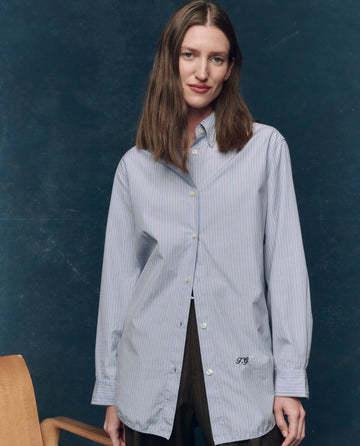 Indoors, a woman with straight brown hair wears THE GREAT. F25 COLLECTORS' The Menswear Button Up -- Alumni Stripe, a light blue striped superfine cotton shirt, paired with dark pants and standing beside a wooden chair.