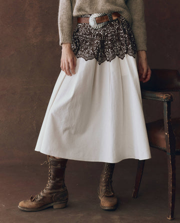 A person wearing THE GREAT. F25 D1's The Regency Skirt in Cream With Western Lace Print, paired with a knit sweater, brown lace-up boots, brown belt with silver buckle, and dark lace overlay stands next to a brown chair.