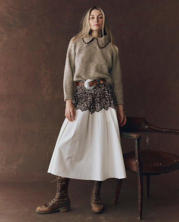 A woman indoors wears THE GREAT. F25 D1's The Crochet Collar Pullover in Oat with Brown, paired with a wide lace-detailed belt, long white skirt, and brown lace-up boots. There’s an empty chair next to her.
