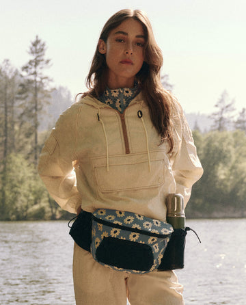 A woman in a beige jacket and floral neck scarf stands by a lake with trees behind her, carrying THE GREAT. SP25 TGO's Adventure Hip Pack in Falling Daisy Multi, complete with an attached thermos.