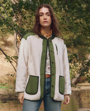 Outdoors, a person wears THE GREAT. F25 TGO's Plush Tundra Jacket in Cream & Army—plush fleece with multiple pockets—paired with a patterned scarf and jeans, surrounded by trees and greenery.