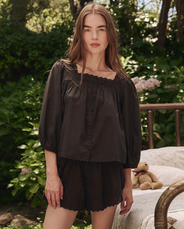 A woman with long brown hair stands outdoors wearing THE GREAT. SP25 SLEEP's The Eyelet Button Sleep Top in black, featuring Victorian-inspired eyelet lace trim and matching shorts. Green foliage and a teddy bear on a bench are in the background.