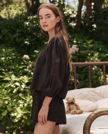 A woman with long brown hair wears THE GREAT. SP25 SLEEP The Eyelet Tap Short in black, standing outdoors near a bed with greenery and a teddy bear in the background.