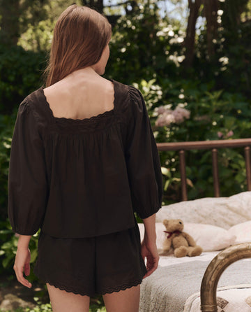 A woman with long brown hair wears The Eyelet Button Sleep Top in black by THE GREAT. SP25 SLEEP, standing outdoors near a brass bed with a floral quilt and teddy bear. Victorian-inspired eyelet lace trim adds charm to her look.