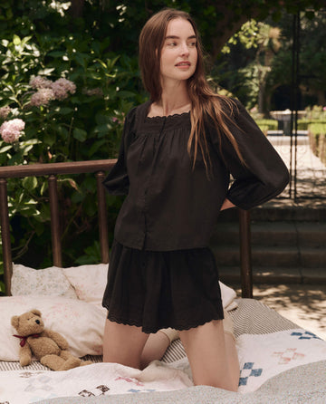 A woman wearing THE GREAT. SP25 SLEEP's black Eyelet Tap Short kneels on a bed outdoors, with a teddy bear and patterned quilts beside her, surrounded by greenery and flowers.