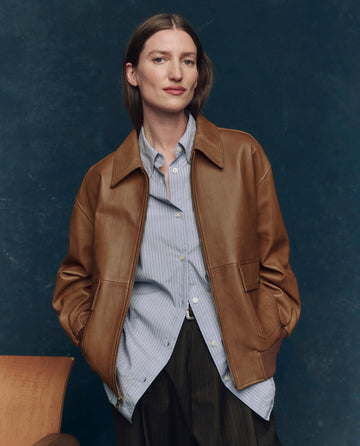 A person stands with hands in the pockets of THE GREAT. F25 COLLECTORS' "The Flight Jacket" in Saddle Brown, styled with a blue striped shirt and dark pants, posed against a dark blue background.