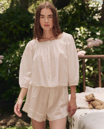 A woman in THE GREAT. SP25 SLEEP's True White Eyelet Tap Short stands outdoors by a bed with a teddy bear, surrounded by greenery and flowers.