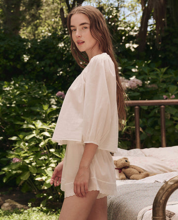 A woman with long hair stands outdoors amid lush greenery and flowers, wearing THE GREAT. SP25 SLEEP The Eyelet Button Sleep Top in True White cotton voile, near a bed with a teddy bear for a dreamy scene.