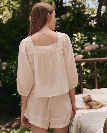 A woman with light brown hair stands outdoors, facing away, wearing THE GREAT. SP25 SLEEP The Eyelet Tap Short in True White. Behind her, a teddy bear and blanket rest on a bed surrounded by greenery.