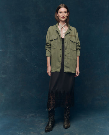 A woman stands against a dark blue backdrop wearing THE GREAT. SP25 COLLECTORS The Vintage Army Jacket -- Classic Army over a black dress with lace trim and black cowboy boots.