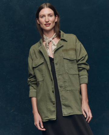A woman stands against a dark background wearing THE GREAT. SP25 COLLECTORS The Vintage Army Jacket -- Classic Army in cotton slub twill over a black dress, accessorized with a light scarf around her neck.