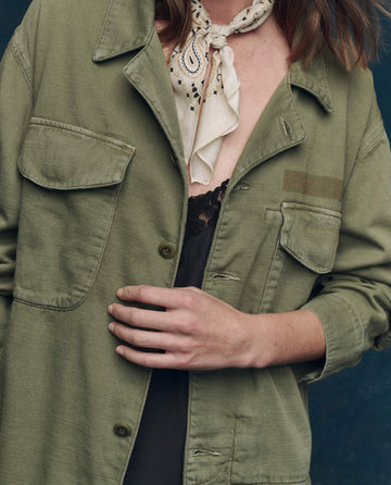 A person wears The Vintage Army Jacket – Classic Army by THE GREAT. SP25 COLLECTORS in cotton slub twill over a black lace top, accessorized with a patterned neck scarf.