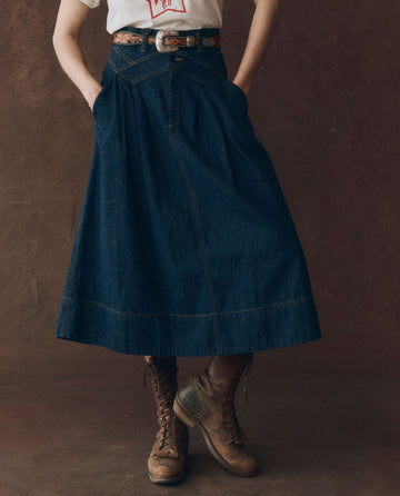 Person wearing a white shirt, THE GREAT. F25 D1 The Field Skirt in Midnight Rinse with a brown belt and brown lace-up boots, standing against a plain dark background.