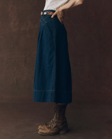 Person standing sideways wearing a white shirt, The Field Skirt – Midnight Rinse by THE GREAT. F25 D1, brown belt, and worn brown lace-up boots against a brown background.