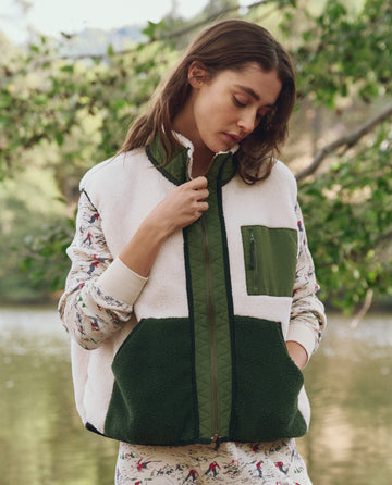A woman stands by the water in THE GREAT. F25 TGO’s Plush Tundra Vest—Cream & Army—with a zip and large pockets, worn over a printed shirt, inspired by vintage camping gear, green foliage behind her.