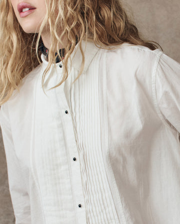 A person with wavy blonde hair is shown from the shoulders up, wearing THE GREAT. H25 COLLECTION's The Classic Tux Top.. in white, featuring pleated detailing.