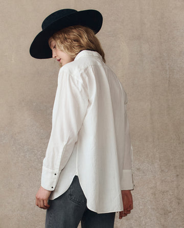 Model wears THE GREAT. H25 COLLECTION's Classic Tux Top in white, paired with gray jeans and a black wide-brimmed hat, standing sideways against a neutral background.