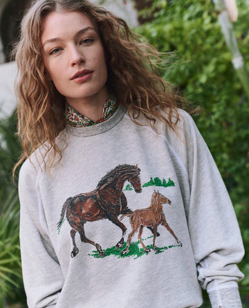 A woman with wavy hair wears THE GREAT. PF25’s The College Sweatshirt in soft heather grey with a running horses design, standing outdoors amid greenery.