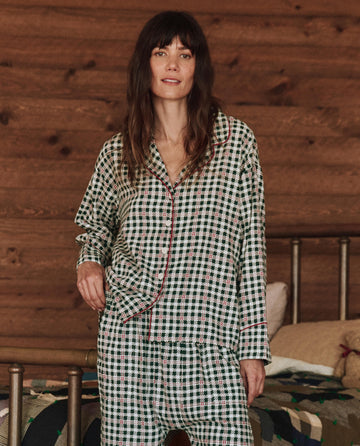 A person wearing The Pajama Shirt in Winter Pine Plaid by THE GREAT. HOL 24 SLEEP stands in a cozy wooden room with a bed.