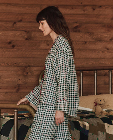 A person wearing The Pajama Shirt in Winter Pine Plaid by THE GREAT. HOL 24 SLEEP stands by a bed decorated with a patchwork quilt and a teddy bear, set against a wooden wall backdrop.