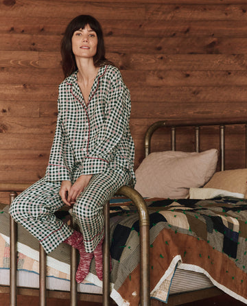 A person in **THE GREAT. HOL 24 SLEEP**'s **The Pajama Shirt - Winter Pine Plaid** sits on a bed with a quilt in a room with wooden walls, their checkered pajama shirt perfectly complementing the relaxed ambiance.