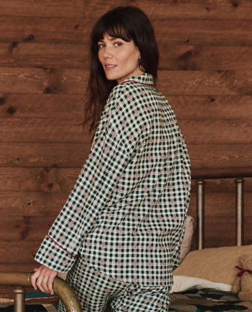 A person in "The Pajama Shirt - Winter Pine Plaid" by THE GREAT. HOL 24 SLEEP sits on a bed with a wooden headboard, framed by a warm wood-paneled wall in the background.
