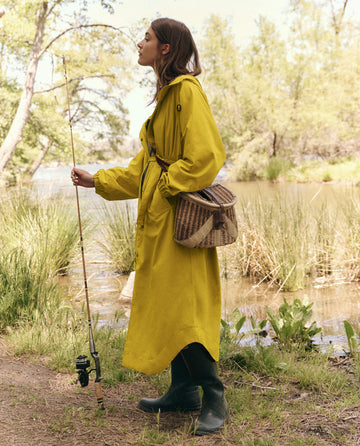 A person wearing 'The Rain Trench -- Sunshine' by THE GREAT. SP25 TGO stands by a pond with a fishing rod and brown bag over their shoulder, surrounded by green boots and lush greenery in a peaceful scene.