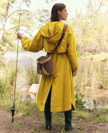 A person in "The Rain Trench" - Sunshine by THE GREAT. SP25 TGO, wearing green boots and holding a fishing rod, stands lakeside with their woven basket over the shoulder in a natural, grassy setting.