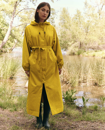 By a lake, a person in a Sunshine Rain Trench by THE GREAT. SP25 TGO and green boots enjoys comfort and dryness amid tranquil trees and shrubs.