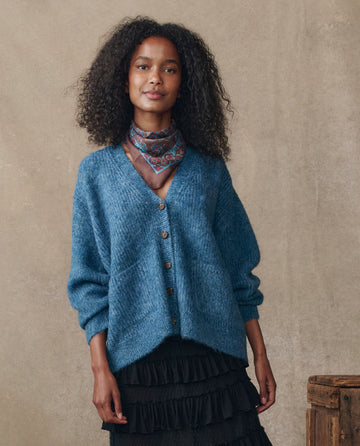 A woman with curly hair wears THE GREAT. H25 COLLECTION The Brushed Melange Cardigan in Wintertide Blue, paired with a patterned scarf and black layered skirt, standing before a neutral textured backdrop.