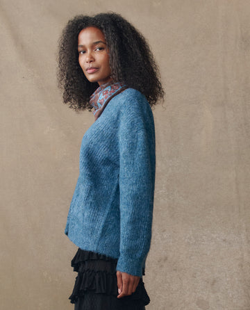 A woman with curly hair stands sideways against a beige background, wearing THE GREAT. H25 COLLECTION's The Brushed Melange Cardigan in Wintertide Blue, paired with a patterned scarf and a black ruffled skirt.