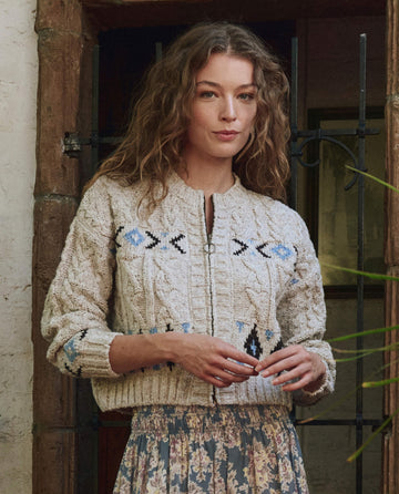 A woman with wavy hair stands by a rustic wooden door, wearing THE GREAT. PF25's The Cavern Cardigan—Oat, a light knit sweater with a geometric pattern—paired with a floral skirt.