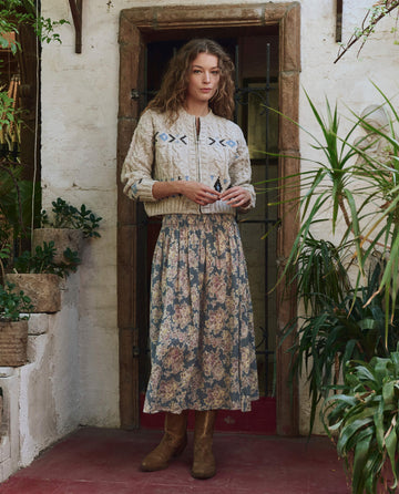 Indoors near plants, a woman wears a cream knit sweater, brown boots, and The Canary Skirt – Indigo Floral by THE GREAT. PF25, featuring a fashionable ankle-length floral design with her hands clasped in front.
