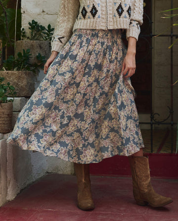 A person wearing a cream knit sweater, brown cowboy boots, and THE GREAT. PF25's The Canary Skirt in Indigo Floral stands on red tiles near potted plants, embodying a unique blend of fashion and charm.