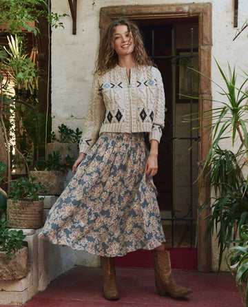 A woman stands indoors wearing THE GREAT. PF25 The Cavern Cardigan in Oat with a long floral skirt and brown boots, surrounded by potted plants and rustic decor.
