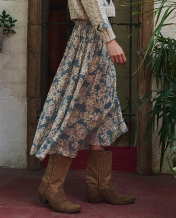 A person in The Canary Skirt – Indigo Floral by THE GREAT. PF25, paired with a knit sweater and brown cowboy boots, stands on a red floor near a rustic door and green plants, highlighting distinctive style.