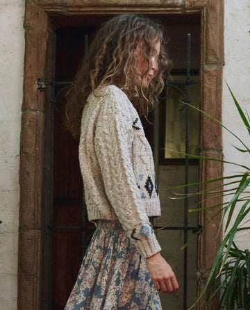 A woman with curly hair stands sideways by a rustic door, wearing a floral skirt and The Cavern Cardigan in Oat from THE GREAT. PF25, her knitwear adding warmth to the scene.