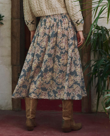 A person wears the Indigo Floral Canary Skirt by THE GREAT. PF25 with a cream knit sweater and tan knee-high boots, standing indoors near plants—a perfect mix of style and comfort.