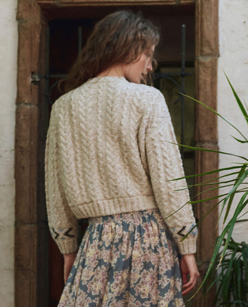 A woman with wavy hair faces away, wearing THE GREAT. PF25 The Cavern Cardigan in Oat and a floral skirt, standing before a wooden door with metal bars and an indoor plant nearby.