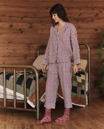 A person wearing The Pajama Shirt in Bordeaux Plaid by THE GREAT. HOL 24 SLEEP, along with cozy socks, stands by a bed adorned with a patchwork quilt, enclosed within a warm wooden room.