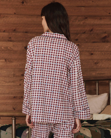 A person in "The Pajama Shirt" by THE GREAT. HOL 24 SLEEP, styled in a classic Bordeaux Plaid, stands with their back to the camera in a cozy, wood-paneled room.