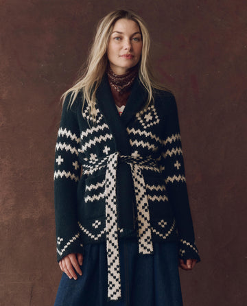 A woman stands against a brown background, wearing THE GREAT. F25 D1 The Reversible Shepherd Cardigan in navy and cream with geometric patterns and a matching belt, layered over a long dark skirt and patterned scarf.