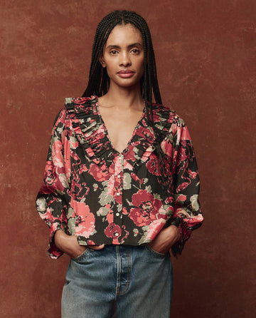 A person stands with hands in their jean pockets, wearing The Symphony Top—Charming Rose by THE GREAT. HOL 24—a floral blouse featuring ruffled details—against a brown textured background.
