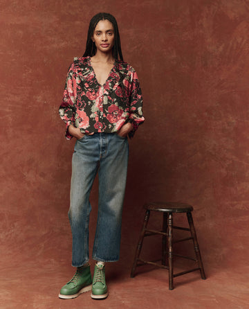 A person wearing The Symphony Top in Charming Rose by THE GREAT. HOL 24, complemented by jeans and green boots, stands gracefully beside a wooden stool against a brown backdrop.