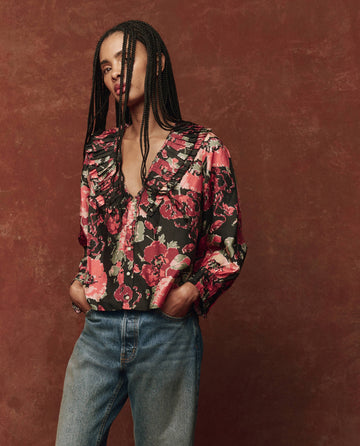 A person with long braided hair wearing The Symphony Top in Charming Rose by THE GREAT. HOL 24 and jeans stands against a brown textured background.